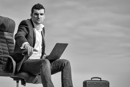 handsome male businessman with courageous face in black formal jacket and white shirt working on laptop sitting on leather office arm chairの写真素材