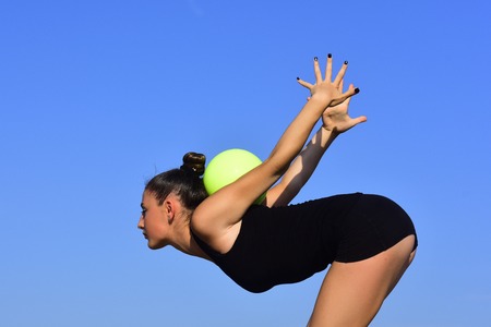 Gymnastic school and energy. Woman gymnast in black sportswear with green ball. Workout of girl on blue sky background. Flexibility in acrobatics and fitness health. Sport and success.の写真素材