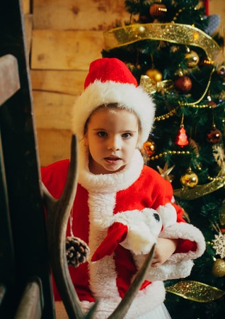 Santa claus kid at Christmas tree. Xmas party celebration. New year small girl in hat. Winter holiday and vacation. Christmas happy child with toy.の写真素材