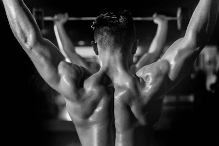 Handsome young man with sexy muscular wet body and bare back training with heavy exercise equipment in gymの写真素材