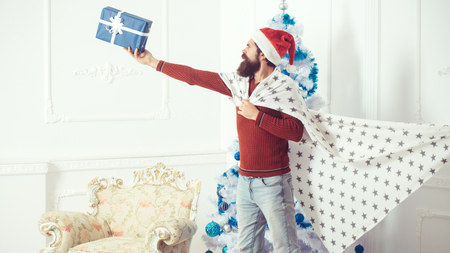 Winter holiday and xmas. Christmas man with beard on serious face hold present box. Santa claus man with toy gift in plaid. New year guy at blue Christmas tree. Boxing day and party celebration.の写真素材