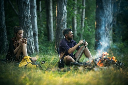 Girl or woman use mobile phone. Man hipster sharpen axe. New technology or archaism concept. Camping, hiking, summer vacation. Couple of hikers relax at bonfire in forest.の写真素材