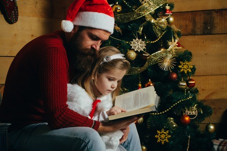New year small girl and man, fairytale. Winter holiday and vacation. Christmas happy child and father read book. Xmas party celebration, fathers day. Santa claus kid and bearded man at Christmas tree.の写真素材