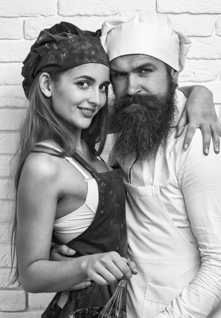 Bearded man, handsome cook or baker and pretty girl or beautiful woman in red apron messy with flour and whisk in bowl hug on white kitchen wallの写真素材