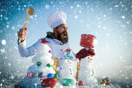 christmas new year snow concept Excited cook, snowmen and snow xmas tree on blue sky. Hipster holding spoon and red pot. Cooking and diet concept. Christmas and new year holidays food. Man in chef hatの写真素材