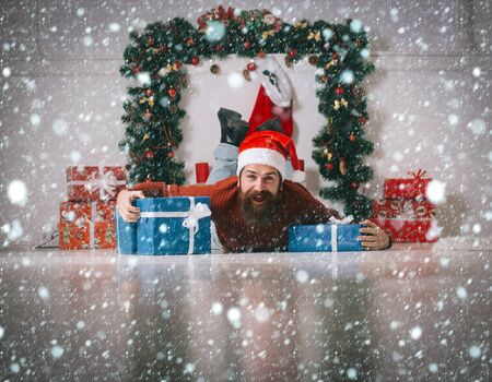 christmas new year snow concept christmas man with beard on happy face in red santa claus hat near present box at new year decoration fireplace, winter holiday celebration, boxing dayの写真素材