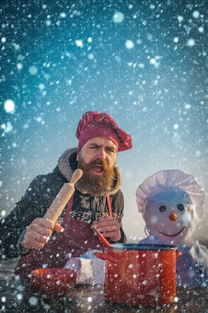 christmas new year snow concept New year guy with kitchen pot. Santa claus man with snowman in hat. xmas leisure and cooking food. Christmas man with beard on happy face. Snowman, winter holidayの写真素材