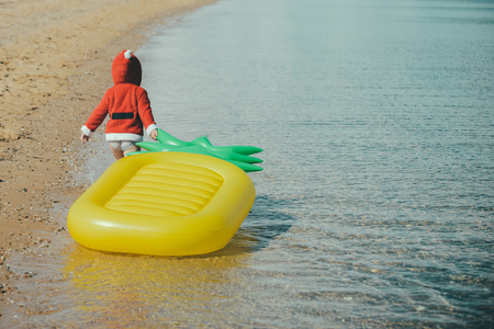 Christmas happy child with pineapple mattress in water. Santa claus kid in red Christmas costume. Winter holiday vacation. Xmas party celebration, childhood. New year small boy at beach.の写真素材