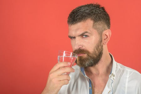 Man with long beard hold water glass on red background. Hipster drink clean healthy water, refreshing. Health and dieting. Hangover and thirst. Life source and healthcare, copy spaceの写真素材