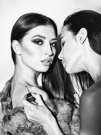 Pretty sexy cute woman in gray fur fashion makeup and girl with black hair paints pink lipstick on body on red studio backgroundの写真素材