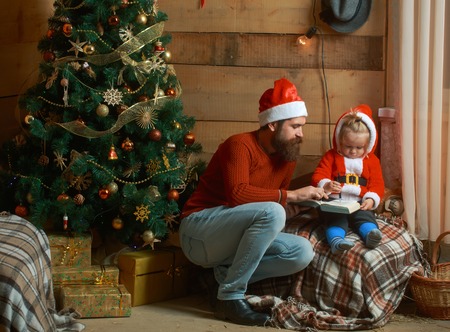 Winter holiday and vacation. Xmas party celebration, fathers day. New year small boy and man, fairytale. Christmas happy child and father read book. Santa claus kid and bearded man at Christmas tree.の写真素材