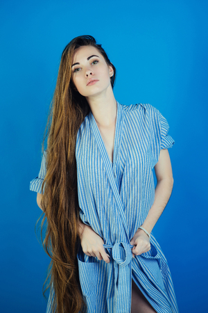 Woman or girl with long hair in striped dressing gown pose on blue background. Beauty salon concept. Look, fashion, style.の写真素材