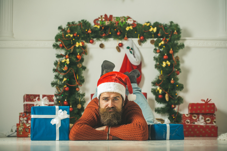 christmas man with beard on sad face in red santa claus hat near present box at new year decoration fireplace, winter holiday celebration, boxing dayの写真素材