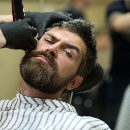 Fashion and beauty, innovation. Haircut of bearded man, archaism. Man cut long beard and mustache with trimmer razor. Barber and hairdresser. Hipster with serious face in barbershop, new technology.の写真素材