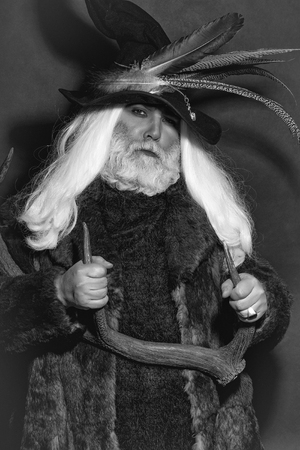 Druid old man with long grey hair beard in hunter hat with bird feathers and fur coat with deer...