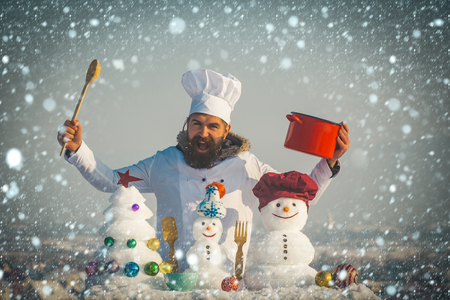 new year christmas snow concept Cook shouting, snowmen and snow xmas tree on blue sky. Cooking and diet concept. Christmas and new year holidays food. Man holding spoon and red pot. Hipster chef hatの写真素材