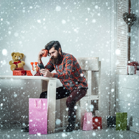 christmas new year snow concept man or hipster, with long beard with smartphone sitting at table with cup, teddy bear, hearts, colorful bags, gift boxes in room on white wall. Valentines dayの写真素材