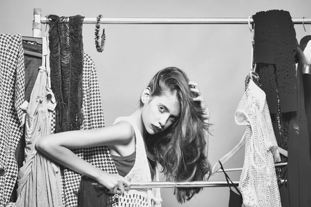 Pretty girl young sexy beautiful woman cute fashion female model with long hair and purple lips among clothes on hangers in wardrobe or shop on grey wall backgroundの写真素材