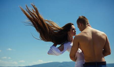 Girl and macho with muscular torso on nature. Couple in love on blue sky. Relationship, romance concept. Man hug woman with flying long hair on sunny day. Summer vacation, wanderlust.の写真素材
