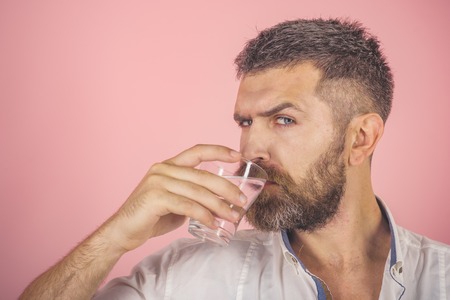 Health and dieting. Hangover and thirst. Life source and healthcare. Man with long beard hold water glass on pink background. Hipster drink clean healthy water, refreshing, copy spaceの写真素材