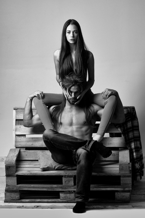 Young couple of pretty girl or cute woman, with brunette long hair and handsome man or muscular macho with muscular torso, sits on wooden, pallet sofa on grey backgroundの写真素材