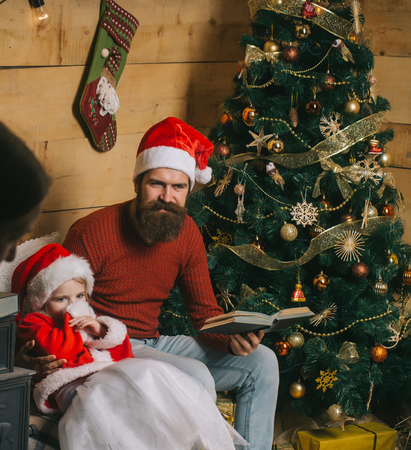 Christmas happy child and father read book. Santa claus kid and bearded man at Christmas tree. Winter holiday and vacation. Xmas party celebration, fathers day. New year small girl and man, fairytale.の写真素材