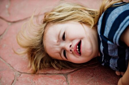 Kid boy with long blond hair cry on brown tile outdoor. Child and childhood concept.の写真素材