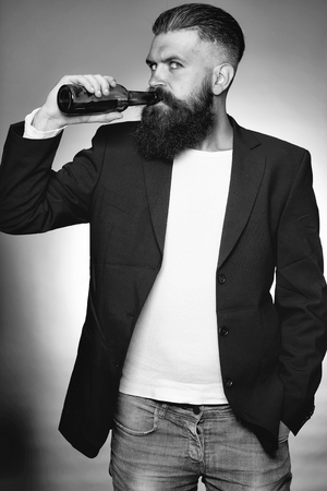 Handsome young man with long beard and moustache in black jacket holding green glass beer bottle in studio on grey backgroundの写真素材