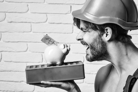 young handsome bearded macho man smiling builder with sexy muscular athletic strong body has strong hands in orange hard hat or helmet holds pig dollar moneybox and brick on white wall backgroundの写真素材