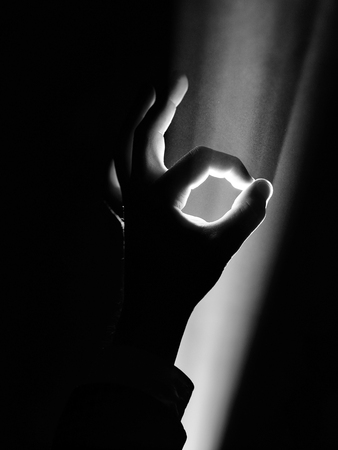 dark silhouette of human male hand with fingers in spotlight or backlight light with okay or ok gesture on black background with dramatic projector shine ray or beamの写真素材