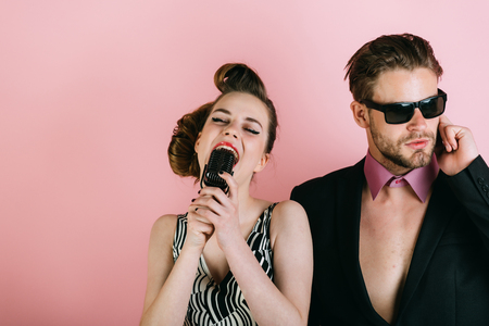 Music and love, security. Pinup girl and man on pink, radio. Beauty, vintage fashion, music band. Singer man and woman with retro hair and makeup. Couple in love in glasses sing in microphone.の写真素材
