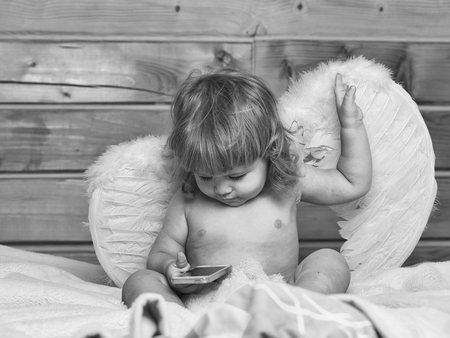 Cute happy beautiful playful child boy with wet hair sitting in hothouse bath white fluffy towel naked indoor on wooden background in feathered angel wings playing on mobile phone, horizontal pictureの写真素材