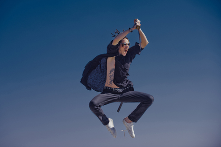 Martial arts, sport activity. Warrior training with katana in sunglasses, black hat and clothes. Concentration and zen concept. Defense, power, honor, force. Man samurai jumping with sword on blue skyの写真素材