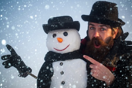 christmas and new year snow concept Man and snowman pointing fingers. Gentlemen in black hats and scarves on blue sky. Surprised hipster with open mouth. Winter holidays celebration. Christmasの写真素材