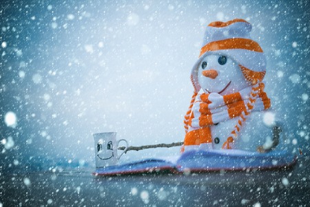 christmas and new year snow concept christmas snowman read book at tea cup with hot wine on table in hat and scarf, copy space, winter holiday celebration, new year and xmasの写真素材