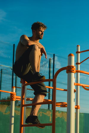 Man sit on metal jungle gym on blue sky. Sportsman with bare torso on monkey bars top. Sport, health, recreation. Climb, climbing frame. Freedom, success, energy concept.の写真素材