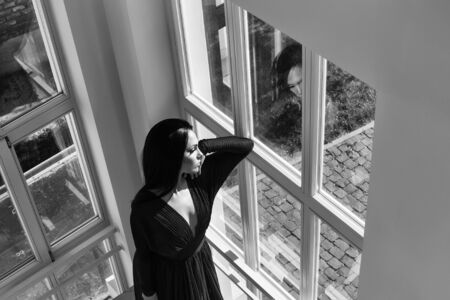 Young beautiful woman with brunette hair in elegant red dress near windowsの写真素材