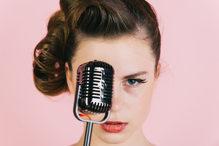 Pin up young girl on pink background, radio. Girl in glasses sing in microphone. Beauty and vintage fashion. Music, look and retro style, pinup. Woman singer with stylish retro hair and makeup.の写真素材