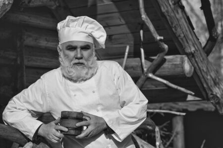 bearded man cook chef in uniform and hat with long beard holding wood cup with iron decoration sunny day outdoor on wooden backgroundの写真素材
