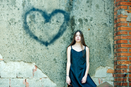 Valentines day holiday celebration concept. Girl posing with heart graffiti on grey wall. Woman wearing black dress on grunge environment. Love symbol of spray paint.の写真素材