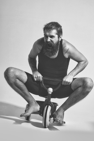 bearded man, long beard, brutal caucasian hipster with moustache in black vest with shouting happy face sitting on colorfyl bicycle toy on grey background, unshaven guyの写真素材