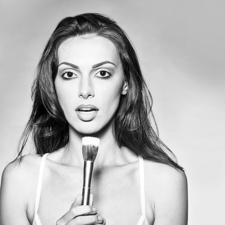 young woman with pretty face and long brunette hair in sexy white bra on body holding fashionable makeup brush set in studio on grey backgroundの写真素材