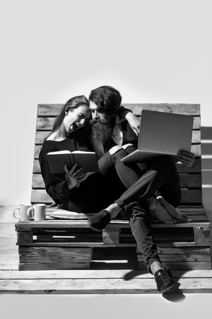 Young couple spend time together on wooden pallet sofa. Handsome man, bearded hipster with beard uses laptop, computer. Pretty winking girl or beautiful woman reads bookの写真素材