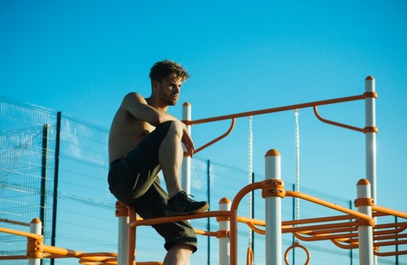 Sportsman with bare torso on monkey bars top. Man sit on metal jungle gym on blue sky. Climb,...