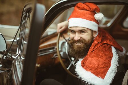 Man in santa hat smile in retro car. Santa Claus driver smiling at wheel. Christmas travel, adventure, journey concept. Merry xmas and happy new year. Winter holidays celebration.の写真素材