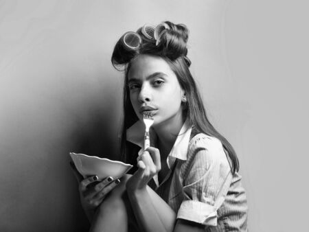Pretty retro girl young sexy cute woman with curlers in long vintage hair in fashionable shirt of maid uniform or housewife on grey background holds white plate and fork ready to eatの写真素材