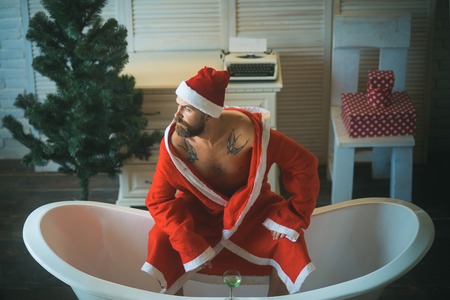 Winter holiday and xmas party. Expectation, love and betrayal. Christmas man with beard, waiting. Home alone, bad santa in bath. Hipster santa at Christmas tree drink wine, loneliness.の写真素材