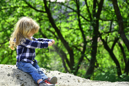 Child play in sand in spring or summer park. Boy sit on pile of sand on idyllic sunny day. Kid with long blond hair in shirt, jeans outdoor. Childhood, playtime concept. Activity, discovery, adventureの写真素材