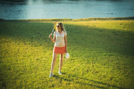 Fashion, beauty, style. Girl athlete in sexy uniform with cap on green grass. Sport, game concept. Activity, energy, health. Woman player with tennis racket walk on lawn.の写真素材