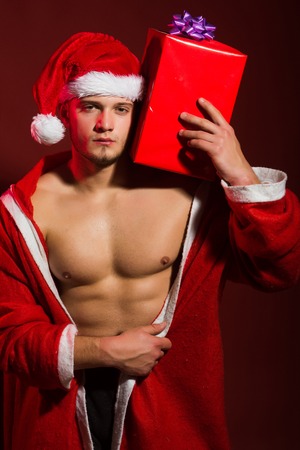 Call boy or sexy athlete man at xmas. Young man in santa costume, present for girls. Christmas party and sex games. New year strip and gift for adult. santa with muscular body in red coat.の写真素材